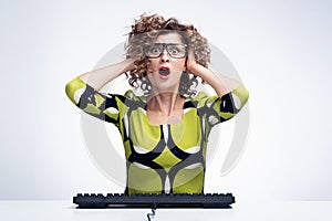 Young emotional scared woman in glasses sitting in front of a computer with a keyboard and clutching her head with her hands