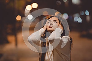 Young emotional woman looking up, city street in the night, evening lights bokeh background outdoors