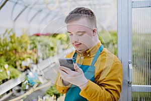 Young employee with Down syndrome working in garden centre, standing in door of greenhouse with cellphone.