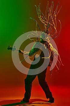 Young energetic musician with long dreadlocks playing electric guitar with passion and energy against gradient red green