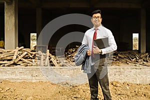 An young engineer in formal shirt and pant with suit and portfolio near a construction site.