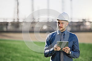 Young engineer with hard hat and tablet standing outdoors by oil refinery.