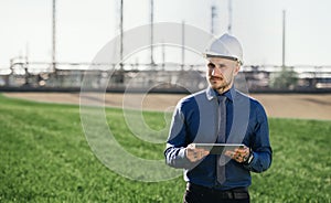 Young engineer with hard hat and tablet standing outdoors by oil refinery.