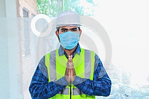 Young engineer wearing mask doing namaste, closeup of beard man wearing blue shirt with yellow vest and white helmet, back to work