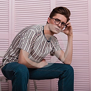 Young Englishman man with a stylish hairstyle in a fashion striped T-shirt in vintage glasses posing on a chair near a wooden pink
