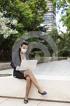 Young enterprising Hispanic woman working with a laptop outdoors. She is wearing a medical mask