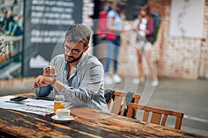 Young entrepreneur businessman with glasses sitting alone at cafe looking at his watch enjoying coffee