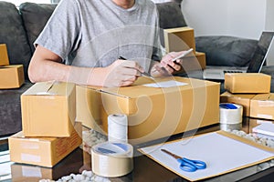 Young entrepreneur SME freelance man using smartphone receive order client and take note working with packaging sort box delivery