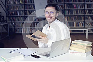 Young entrepreneur or university student smiling, working on laptop reading a book in a library photo