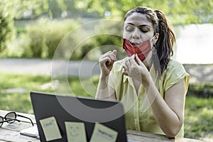Young entrepreneur woman wearing open face mask using laptop outdoors during a video call conference - New normal concept after