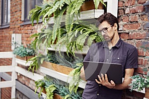 Young entrepreneur working outside using modern laptop. Standing near brick wall, plants, eco office. Successful business people