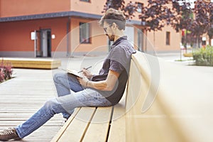 Young entrepreneur working at the park outside on wooden bench. Handsome man wearing glasses using book, writing text.