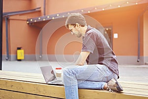 Young entrepreneur working at the park outside on wooden bench. Man using laptop, writing text. Street view. Concept of co-working