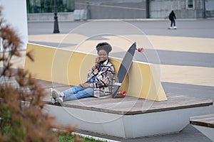Young african girl talk on phone call sit with longboard in urban skate park communicate on mobile