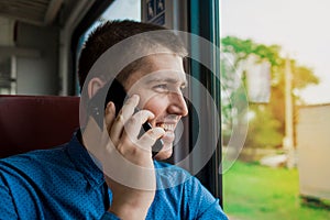 Young european brunette guy in a blue shirt laughs and smiles, looking out the window and talking on a mobile phone in a modern