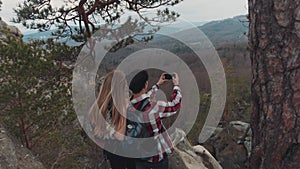 Young European brunette man and blonde girl having a great time together on the top of the mountain, taking photos of