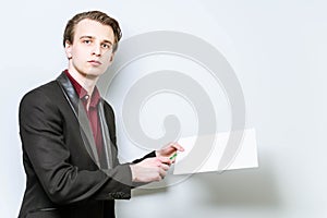 Young european business man holding A4 blank paper and points a finger at it showing data. Business background mockup, with copy