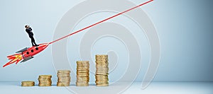 Young european businessman with binoculars standing on flying rocket, growing golden coin chart and mock up place on light wall.