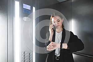 Young european businesswoman is talking on her phone and checking time on her watch in the elevator. Employee using her smartphone