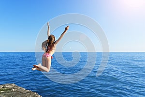 Young european cute girl is jumping and flying to the calm blue sea from concrete breakwater towards summer soft sun