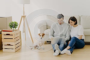 Young European family couple relax during home renovation, focused in laptop computer, plan redecoration, sit near couch with