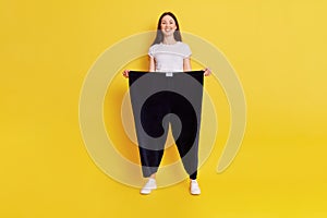 Young European female with dark hair wearing casual white t shirt and black pants too big size, adorable happy woman loose weight