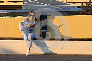 Young european girl chat using smartphone application sitting with longboard skate in park at sunset