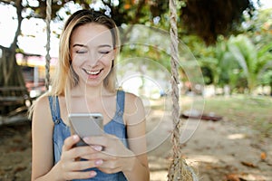 Young european girl riding swing and using smatrphone, sand and tree in background.