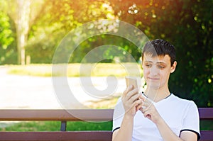 A young european guy sits on a bench in a city park and makes a funny face looking into the phone. The concept of extreme surprise