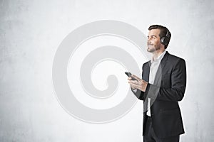 Young European man in elegant suit listening to music with headphones and smartphone, concrete