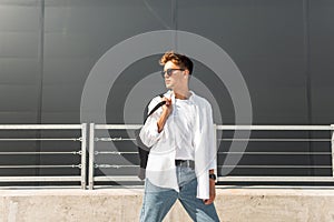 Young European man with a hairstyle in a fashionable shirt in blue jeans in  sunglasses with a bag poses in a modern building