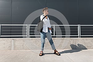 Young European man hipster in a white shirt in blue stylish jeans in dark sunglasses with a black bag stands in the city