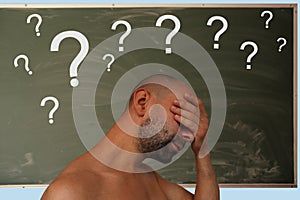 Young European man with a naked torso holding his head against the background of a lot of white question marks on a chalkboard for