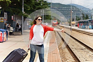 Young european pregnant woman with heavy baggage trying to stop train on railway station for traveling at sunny day