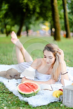 Young european woman lying on plaid in park near watermelon and reaing book.