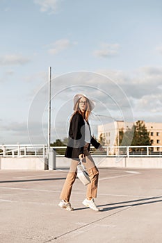 Young European woman model in straw hat in black blazer in fashionable pants in casual sneakers with bag walk outdoors. Lovely