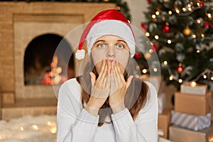 Young European woman wears santa hat and white sweater, being at home around christmas decoration, covers mouth with hands, being