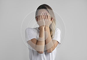 Young european woman in white t-shirt hides her face with both palms, studio photo isolated on gray background. She has social