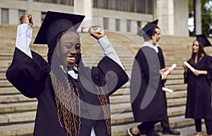 Young excited african american girl student in a university graduate gown and diploma in hands.