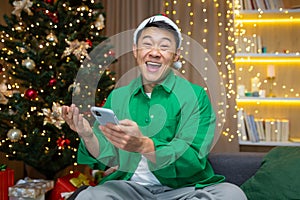Young excited asian man happy positive smile rejoice celebrate victory phone xmas winter sitting at home indoors