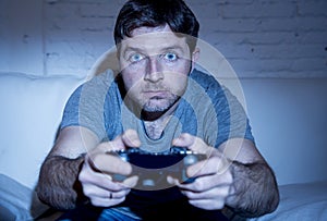 Young excited man at home sitting on living room sofa playing video games using remote control joystick