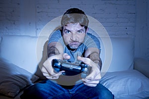 Young excited man at home sitting on living room sofa playing video games using remote control joystick