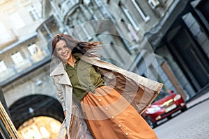 Young excited stylish caucasian woman wearing grey coat looking at camera and smiling while standing on the city street