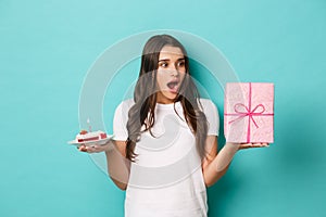 Young excited woman celebrating her birthday, holding bday cake and gift, standing amazed over blue background