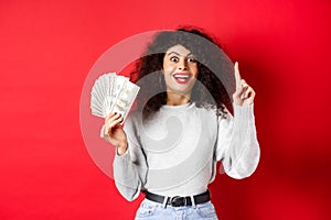 Young excited woman having an idea how make money, showing dollar bills and raising finger in eureka sign, standing on