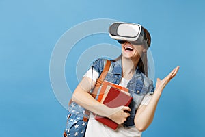 Young excited woman student in denim clothes with backpack wearing virtual reality headset, hold school books spreading