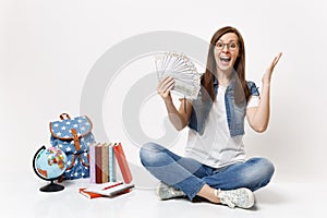 Young excited woman student holding bundle lots of dollars, cash money spread hands sitting near globe backpack, school