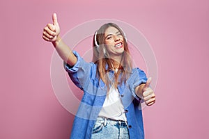 Young excited woman wearing headphones dancing active on pink wall listening to music laughing cheerfully