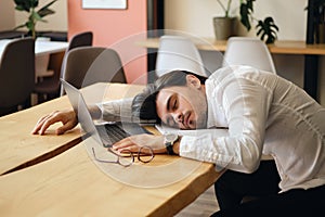 Young exhausted man sitting at the table with laptop while sleeping at work in modern office