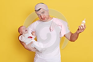 Young exhausted tired father feeding his baby girl with milk bottle isolated over yellow background, poor dad fed up looking after